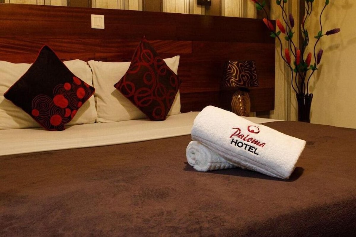 Habitación de hotel con una cama bien hecha, dos almohadas, un florero con flores y una lámpara de pared.