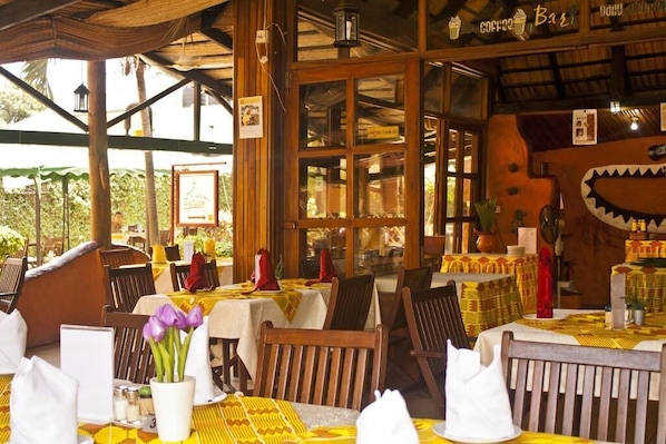 An outdoor restaurant with wooden tables and chairs, covered by a thatched roof, and a sign indicating it serves coffee and BBQ.