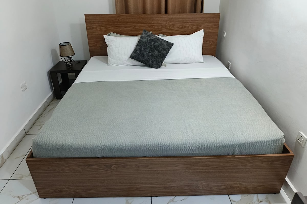 Un lit avec un cadre en bois, deux oreillers, un matelas gris et une petite table de chevet avec une lampe.