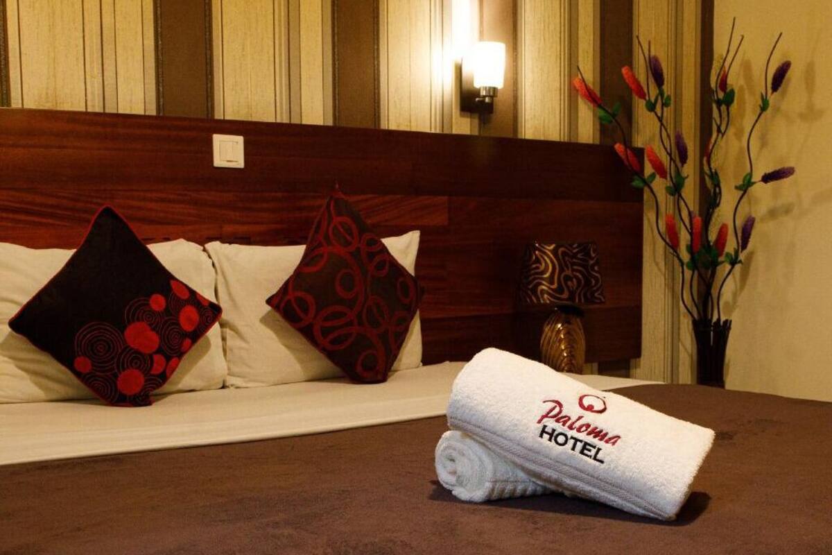 Une chambre d’hôtel avec un lit soigneusement fait, un oreiller à motifs rouges et noirs, et un vase décoratif contenant des branches.