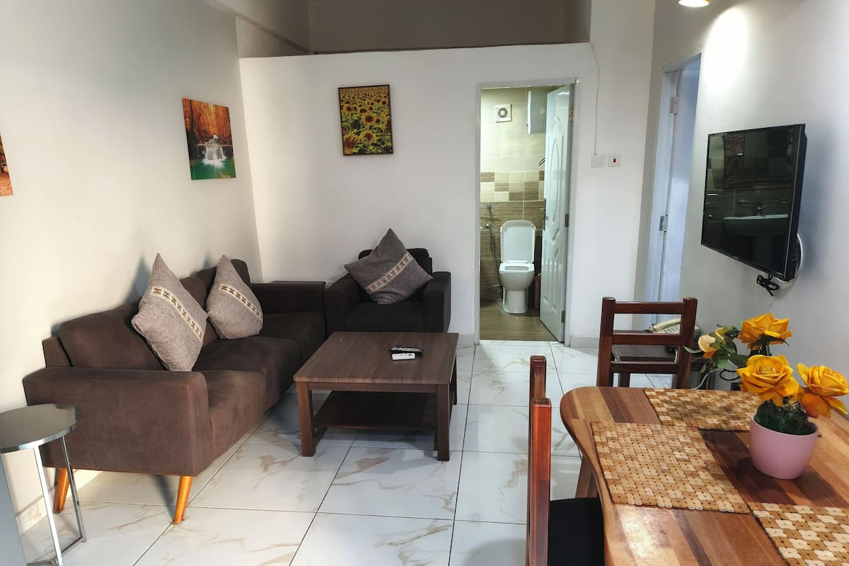 Un salon avec un canapé marron, une table basse, une table à manger avec des chaises et un téléviseur fixé au mur.