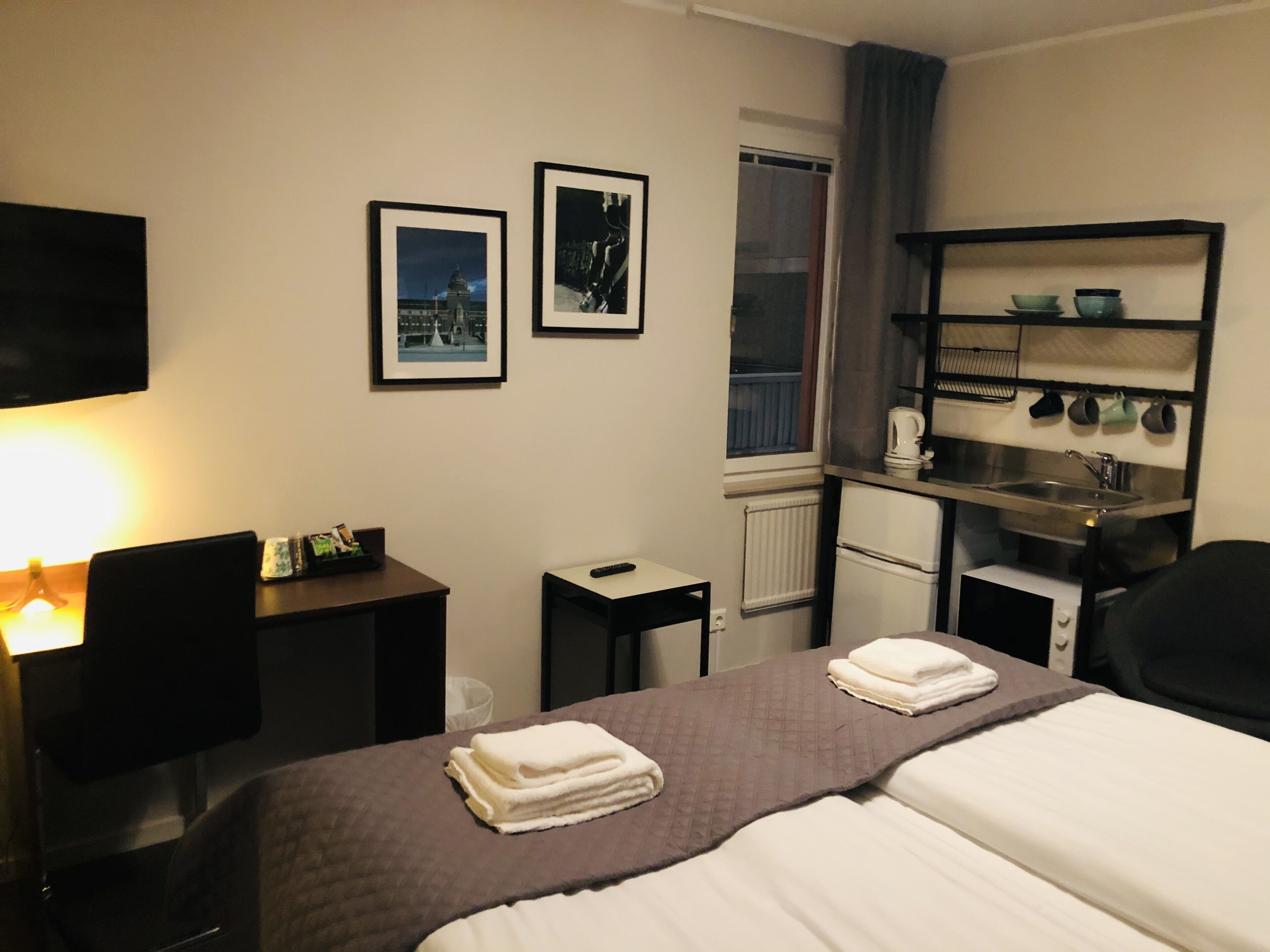 Ett hotellrum med en säng, ett skrivbord, en stol, en tv, en hylla med köksutrustning och två inramade tavlor på väggen.