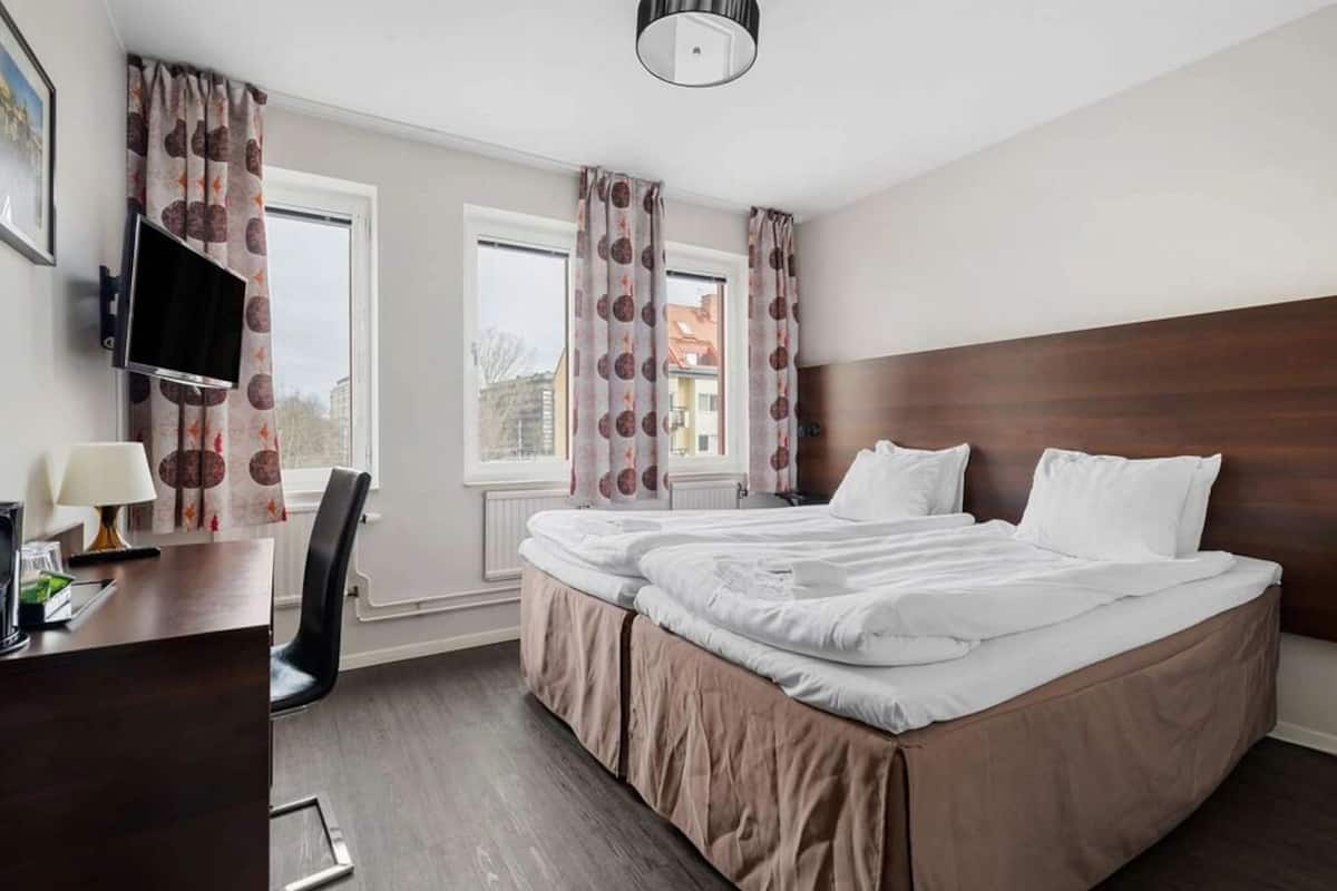 A hotel room with a double bed, a desk with a lamp, a TV mounted on the wall, and two windows with curtains.