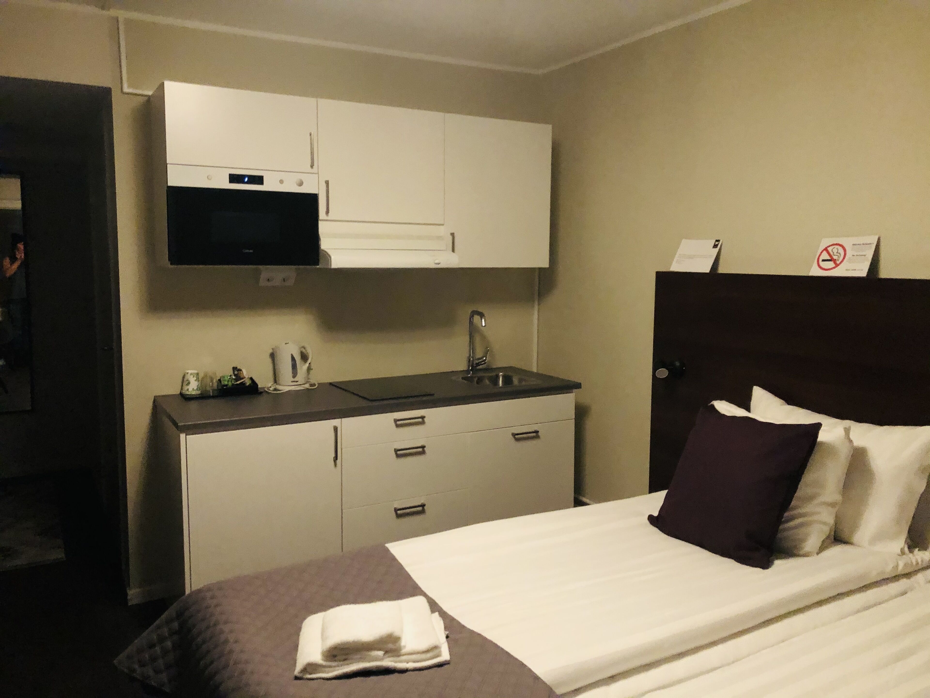 A hotel room with a bed, a small kitchenette with a microwave, a sink, and a cabinet. There is a sign indicating no smoking.