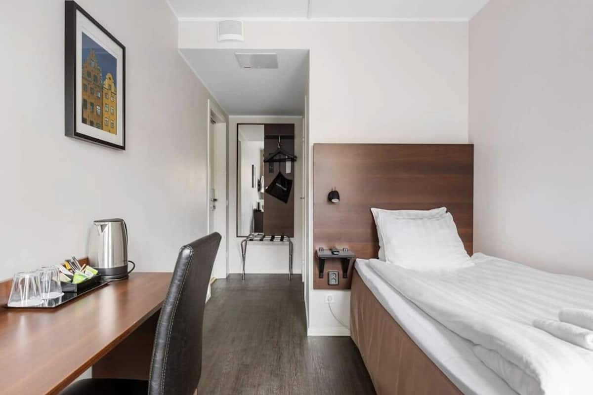 A hotel room with a bed, a desk with a kettle, a chair, and a framed picture on the wall.