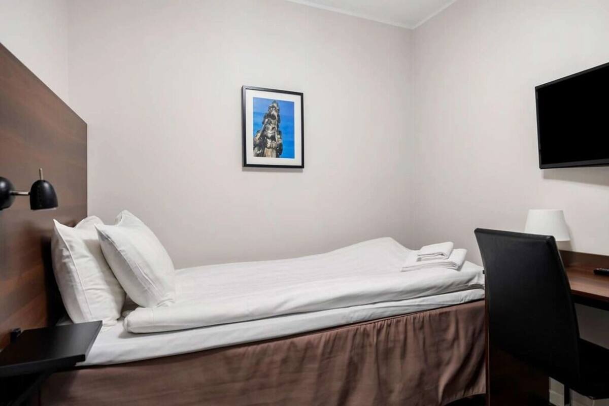 A hotel room with a single bed, a desk, a chair, a flat-screen TV, and a framed picture on the wall.
