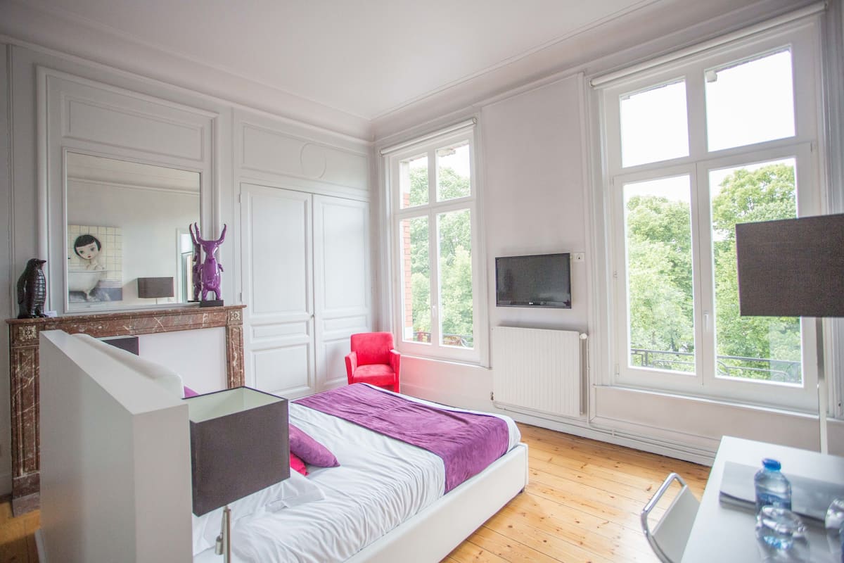 A bedroom with a bed, a red chair, a TV, and large windows.