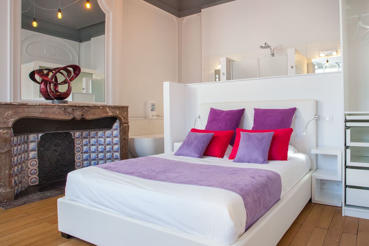 A modern bedroom with a white bed, purple and red pillows, a wooden floor, and a decorative fireplace.