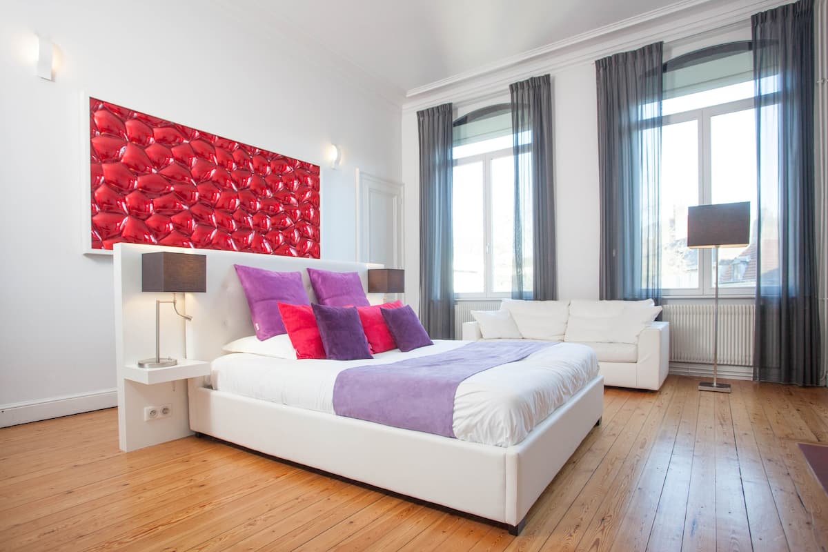 A modern bedroom with a white bed, purple and red pillows, a red textured wall art, a white bedside table, and a white sofa.