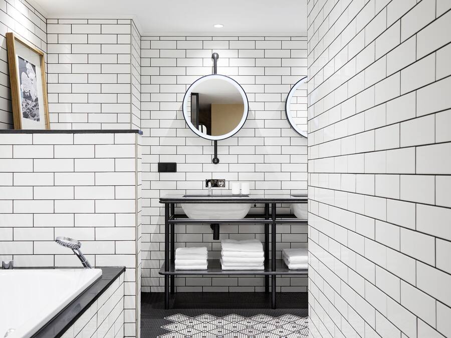 A modern bathroom with white tiled walls, a round mirror, a sink, and a shelf with towels.
