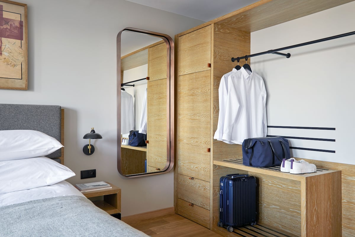 A hotel room with a bed, a bedside table with a lamp, a wardrobe, a mirror, and a bench with a suitcase and shoes.