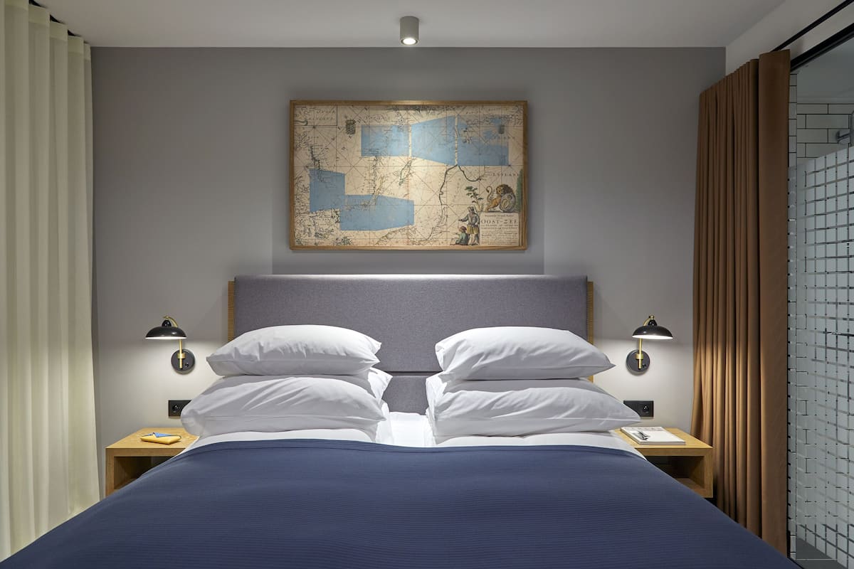 A neatly made bed with white pillows and a blue bedspread, flanked by two bedside tables with lamps, a map on the wall, and a window with curtains.