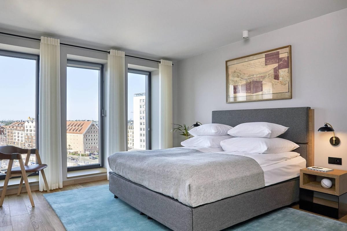 A modern bedroom with a large bed, a chair, a desk, and a city view.