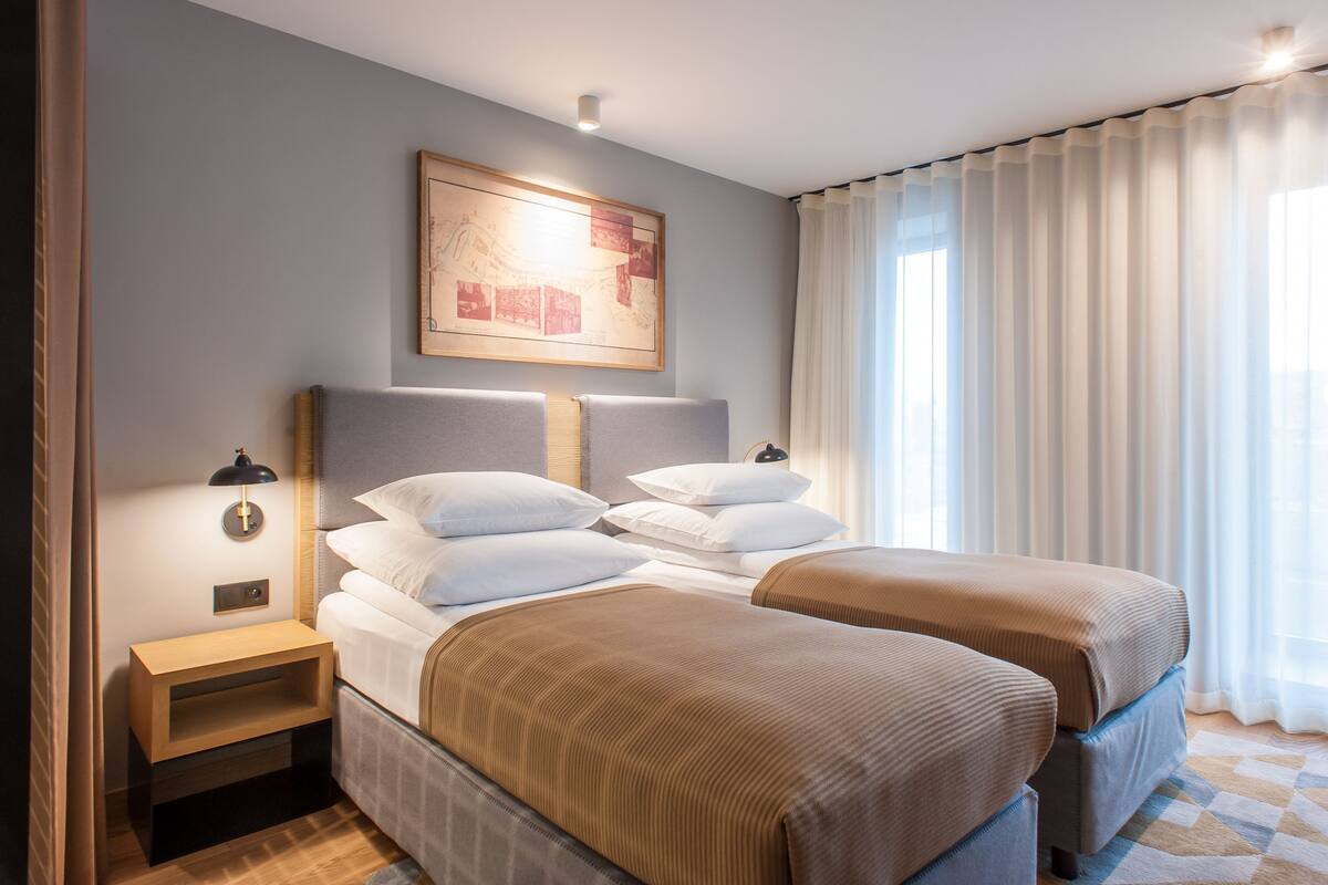 A hotel room with two beds, a nightstand, a wall-mounted lamp, and a large window with curtains.