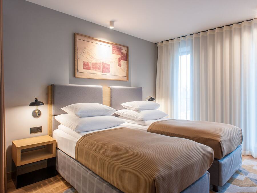 A hotel room with two beds, a nightstand, a wall-mounted lamp, and a large window with curtains.