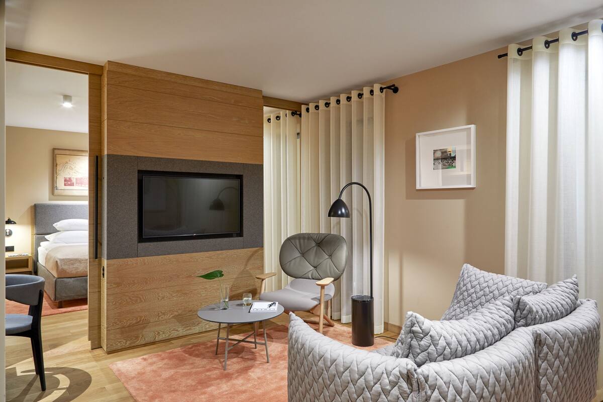 A modern hotel room with a flat-screen TV mounted on a wooden panel, a grey tufted armchair, a round coffee table with a glass and a notebook, a grey sofa, and a large window with sheer curtains.