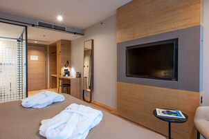 A hotel room with a bed, two white bathrobes, a small table with a magazine, a TV mounted on the wall, a mirror, and a wooden panel design.