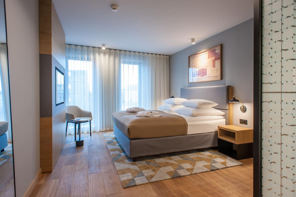 A modern hotel room with a large bed, a desk, a chair, and a view of the city through the window.
