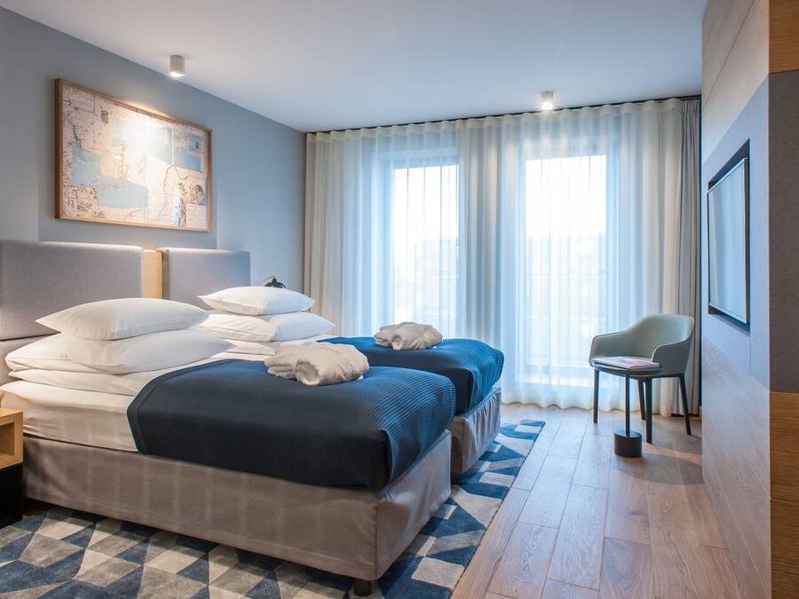 A hotel room with a large bed, two bedside tables, a chair, and a large window with sheer curtains.