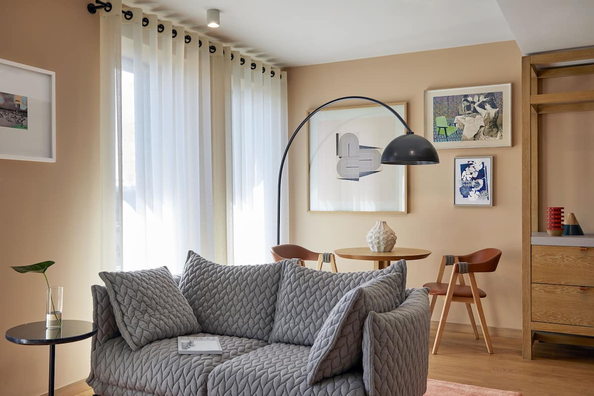 A modern living room with a grey quilted sofa, a small round table, and two chairs. There are framed artworks on the walls and a plant on a side table.