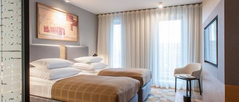 A hotel room with two beds, a sitting area, a large window with curtains, and a framed picture on the wall.