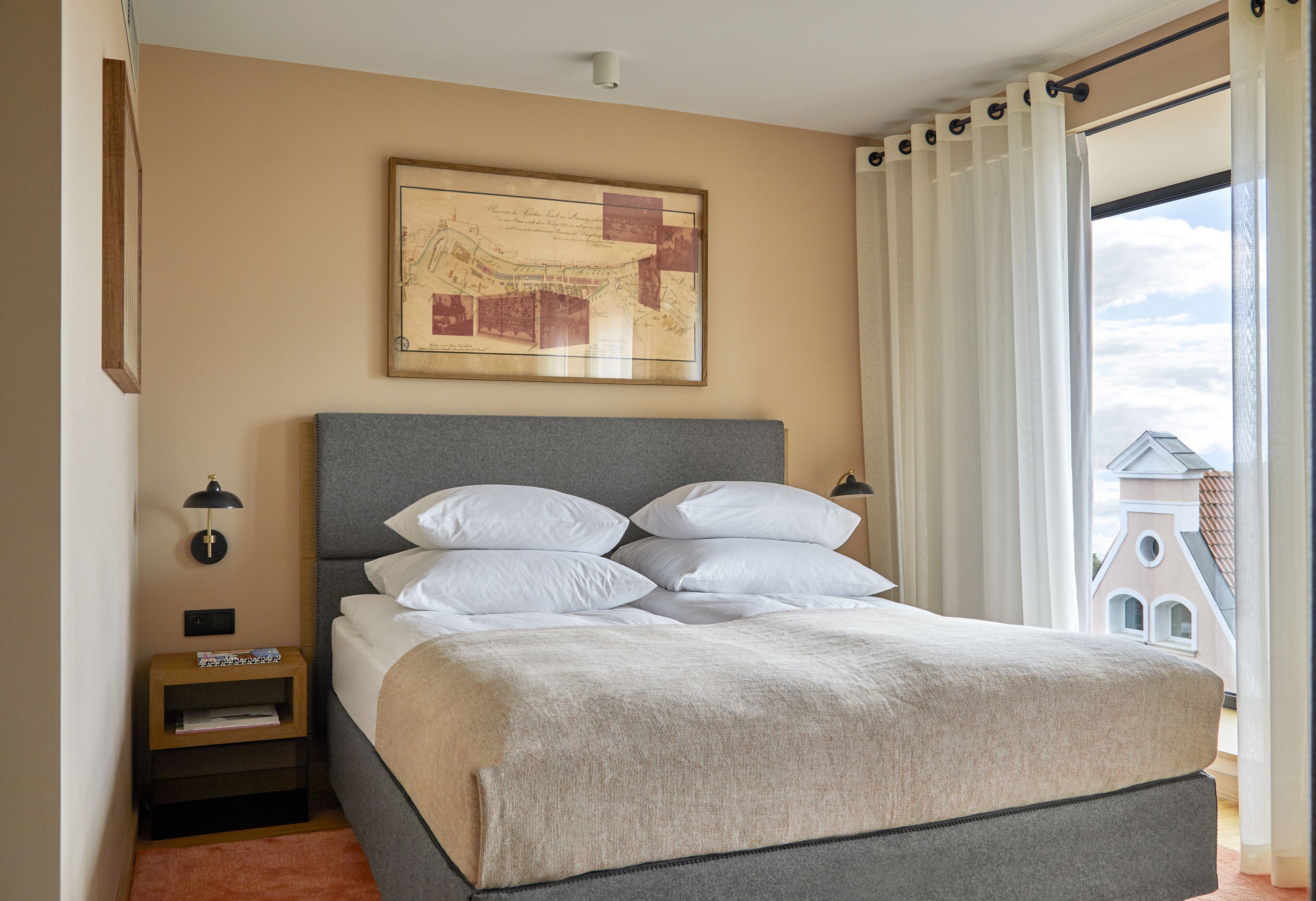 Ein ordentlich bezogenes Bett mit grauem Kopfteil, eine gerahmte Karte an der Wand und Blick auf ein Gebäude durch leichte Vorhänge.