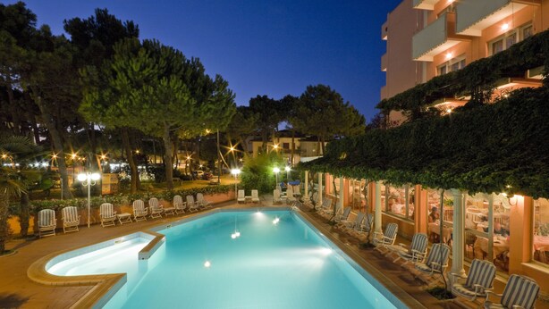 A swimming pool with lounge chairs and a restaurant area at night. - Hotel Meeting (Cesenatico)