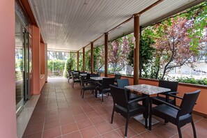 Zona d'un cafè a l'aire lliure amb terra de rajola, mobles de vugada negra i grans finestres que donen vista a arbres i un carrer.