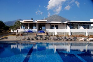 A pool area with lounge chairs and umbrellas, a mountain in the background, and a building with a flat roof. - Samothraki Beach Apartments and Suites Hotel (Samothrace)