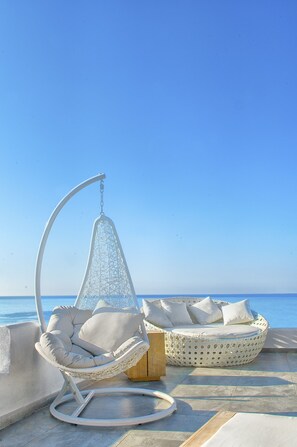 A beachside seating area with a hanging chair and a round cushioned bench, both with white cushions, overlooking the sea. - Samothraki Beach Apartments and Suites Hotel (Samothrace)
