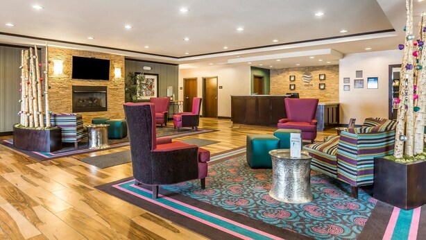 A spacious lobby with a wooden floor, multiple seating areas with colorful chairs, a flat-screen TV mounted on the wall, and a reception desk.