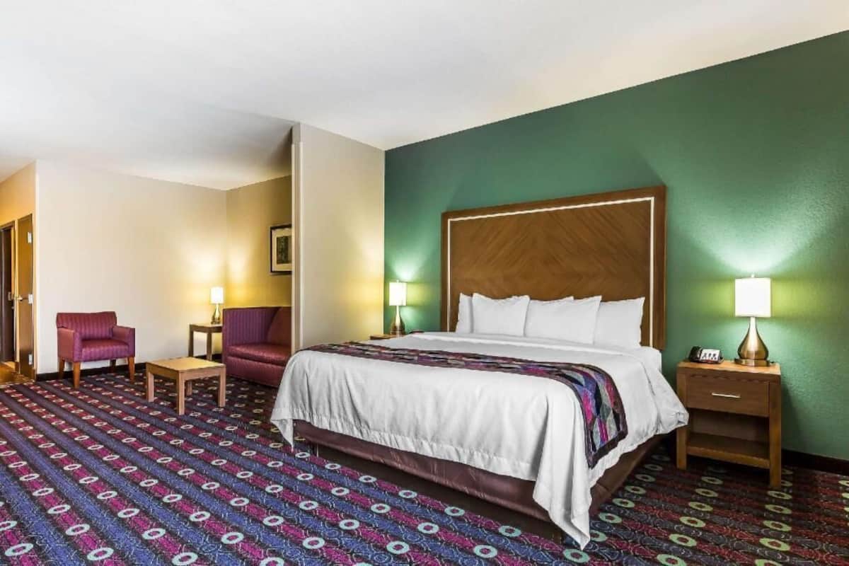 A hotel room with a large bed, two armchairs, a small table, and a nightstand with a lamp.