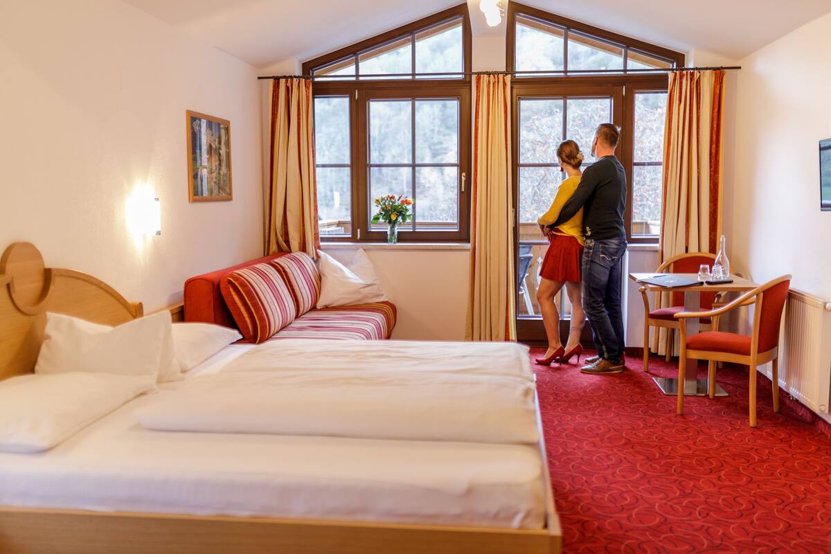 A hotel room with a large bed, a sofa, a dining table with chairs, and a window with curtains.