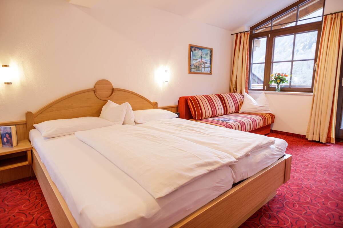 A hotel room with a large bed, a bedside table, a small sofa, a window with curtains, and a painting on the wall.
