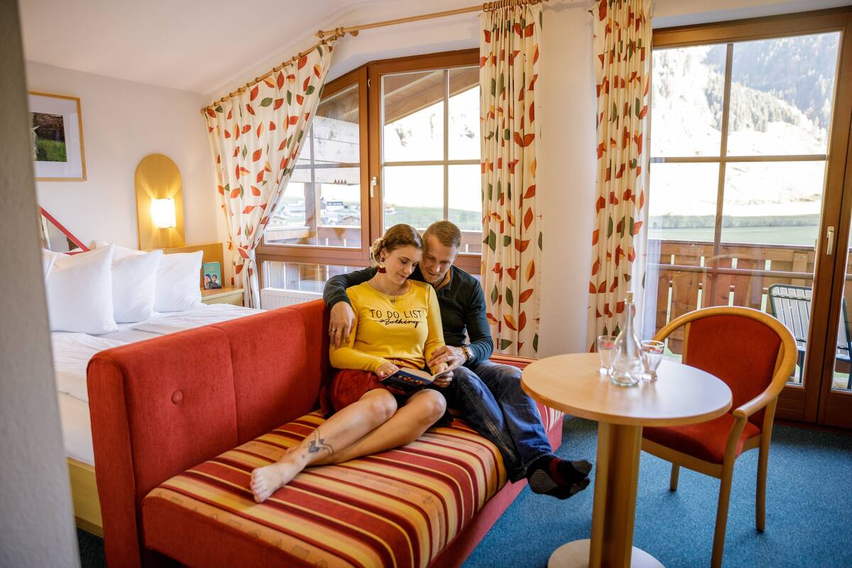 A couple sitting on a red sofa with a view of a mountain landscape through large windows.