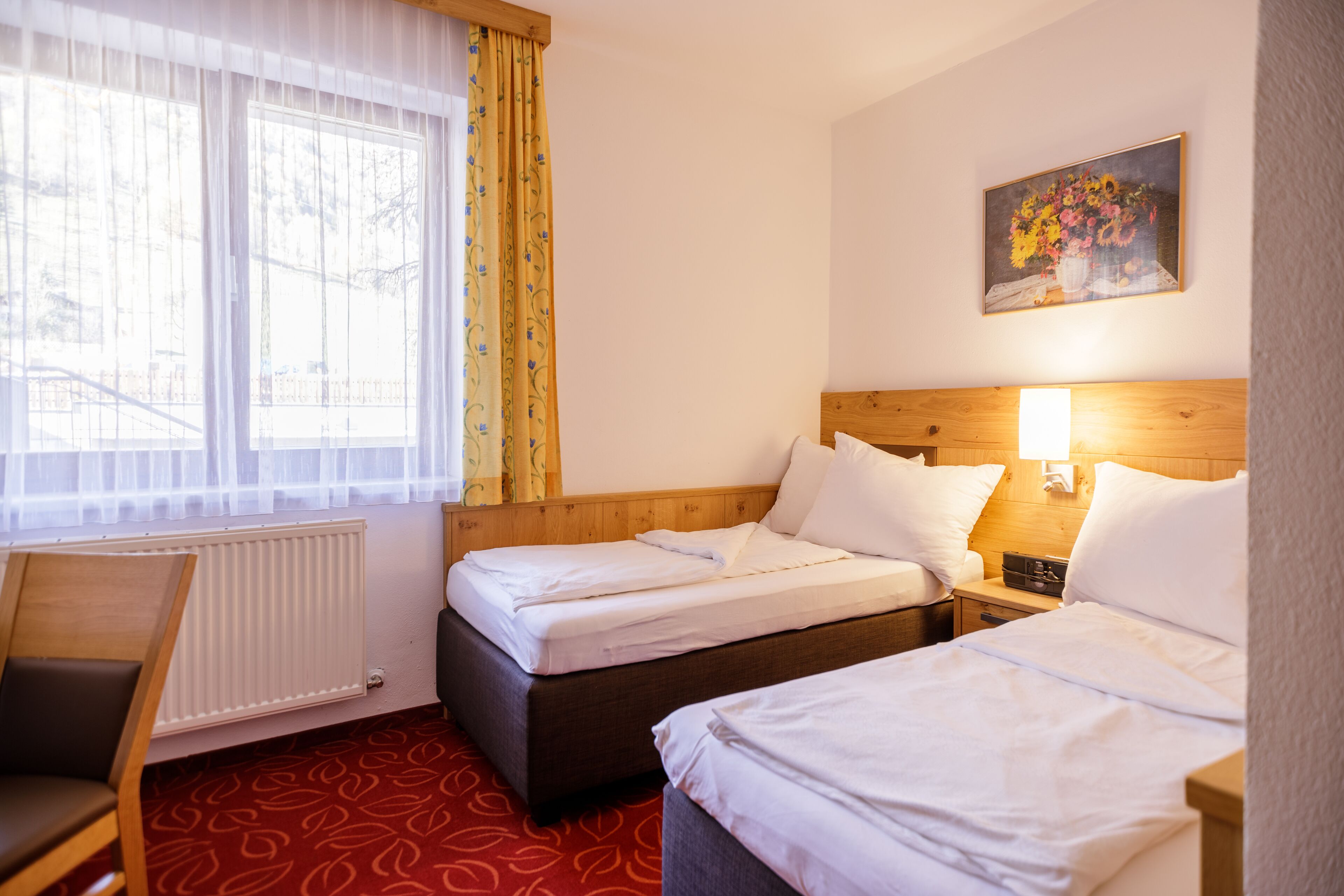 A hotel room with two beds, a wooden headboard, a nightstand with a lamp, a chair, a radiator, and a window with sheer curtains.