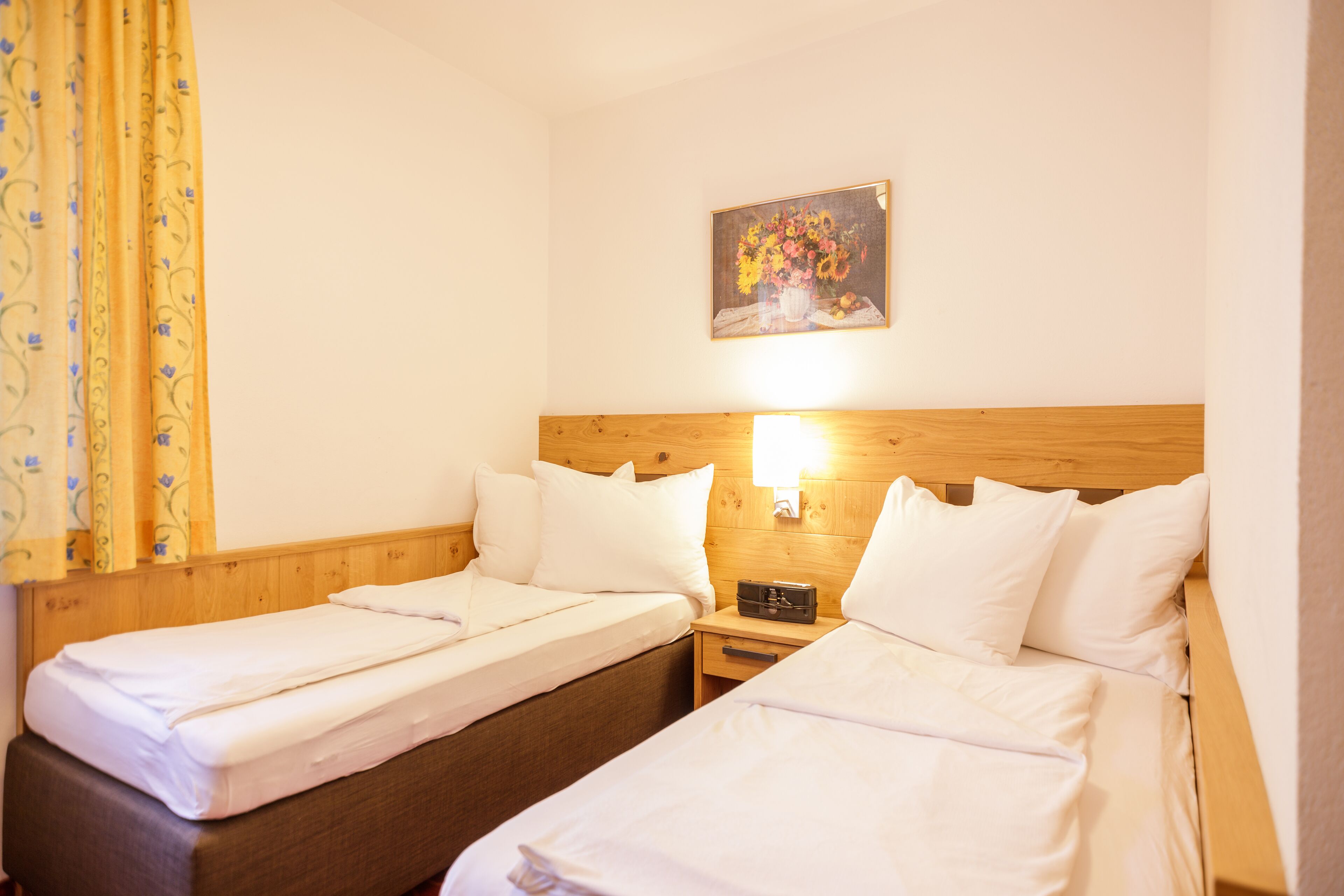 Une chambre d’hôtel avec deux lits simples, des têtes de lit en bois, une table de chevet avec une lampe et un tableau représentant des fleurs accroché au mur.