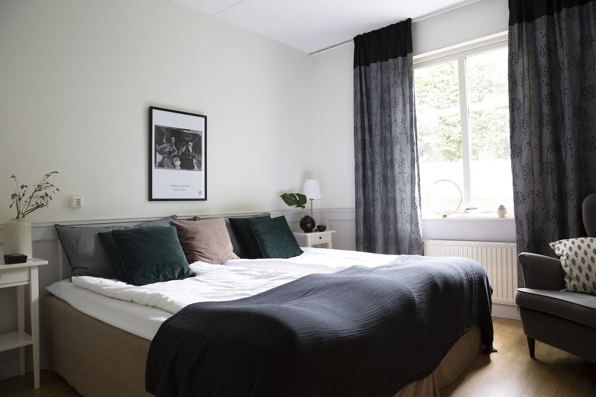 Ein Schlafzimmer mit Bett, Nachttisch mit Lampe, gerahmtem Foto an der Wand und einem Fenster mit Vorhängen.