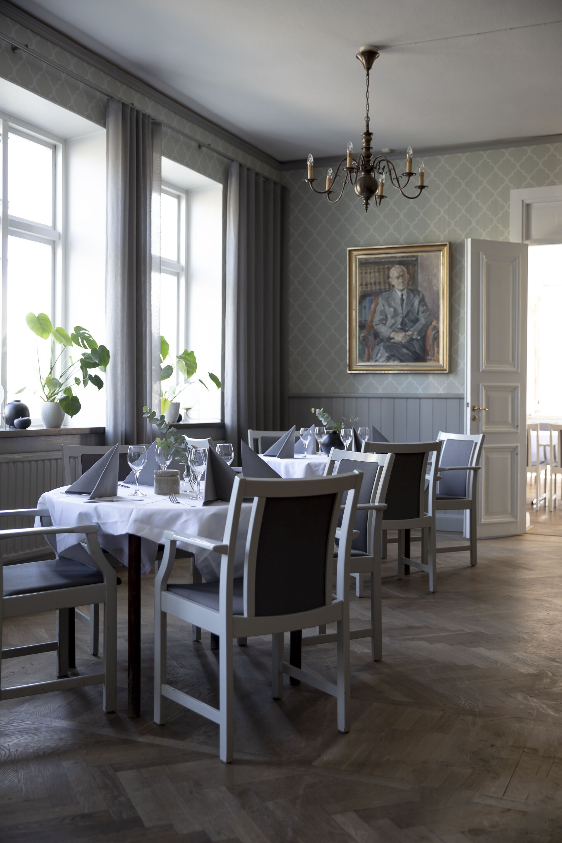 A dining area with tables set for a meal, chairs, a chandelier, and a painting on the wall.