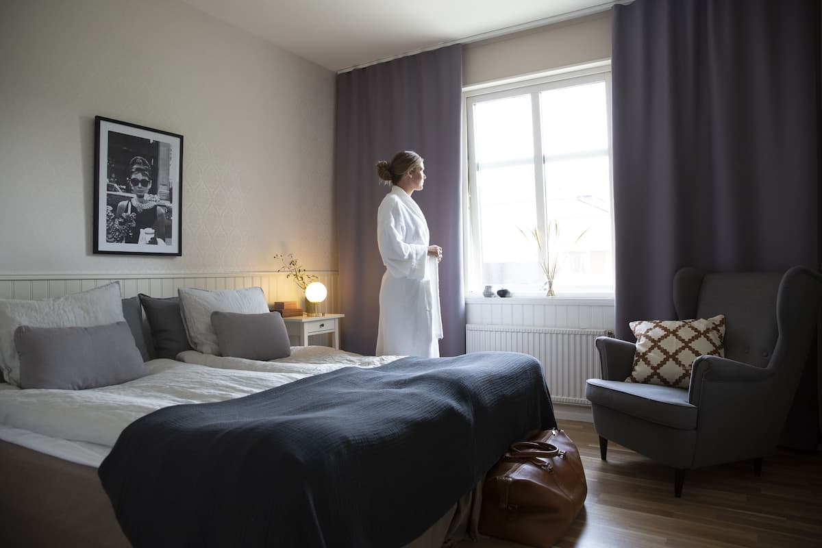 Ein Hotelzimmer mit Bett, Sessel, Nachttisch mit Lampe und einem Fenster mit Vorhängen.