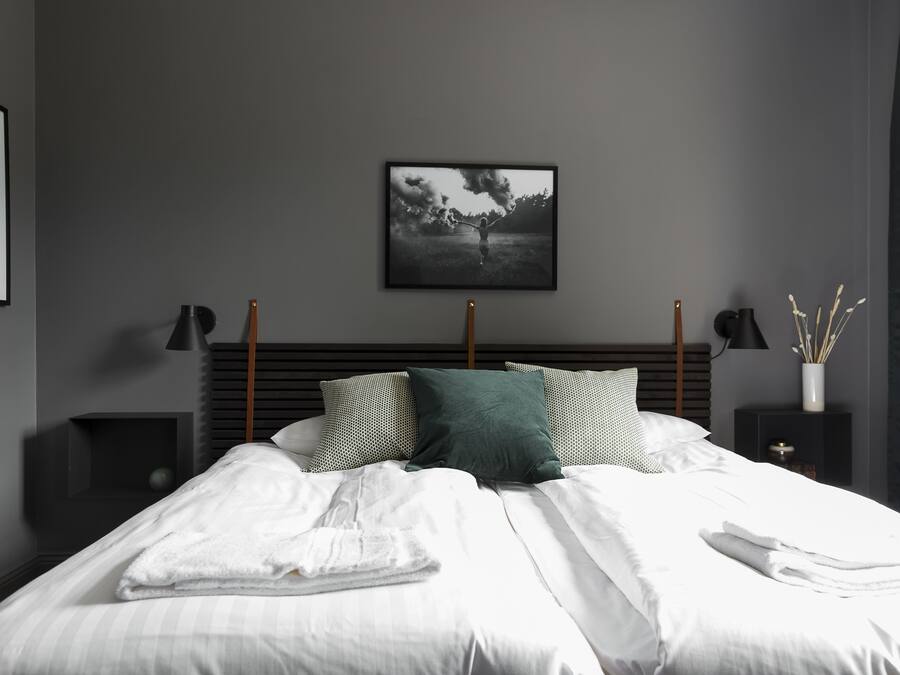 Cama bem arrumada com lençóis brancos e um travesseiro verde, um quadro na parede e duas mesas de cabeceira com abajures.