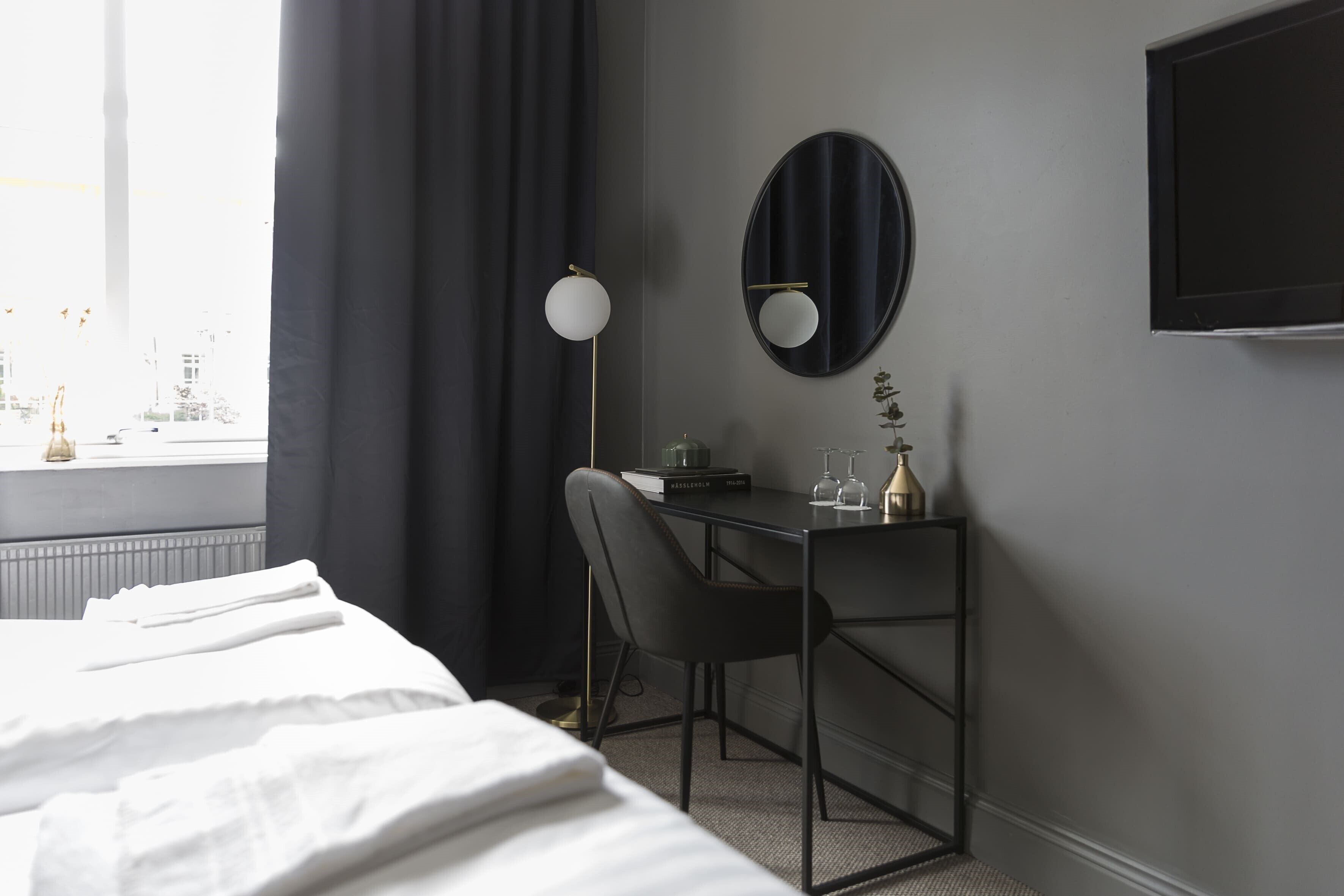 A hotel room with a bed, a desk with a chair, a round mirror, a flat-screen TV, and a window with curtains.