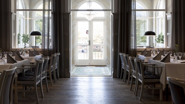 Un comedor formal con mesas dispuestas para el servicio, ventanas amplias con cortinas y un candelabro.