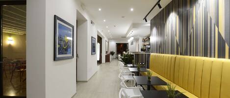 A modern interior with a long dining area, white chairs, black tables, and a yellow cushioned bench.