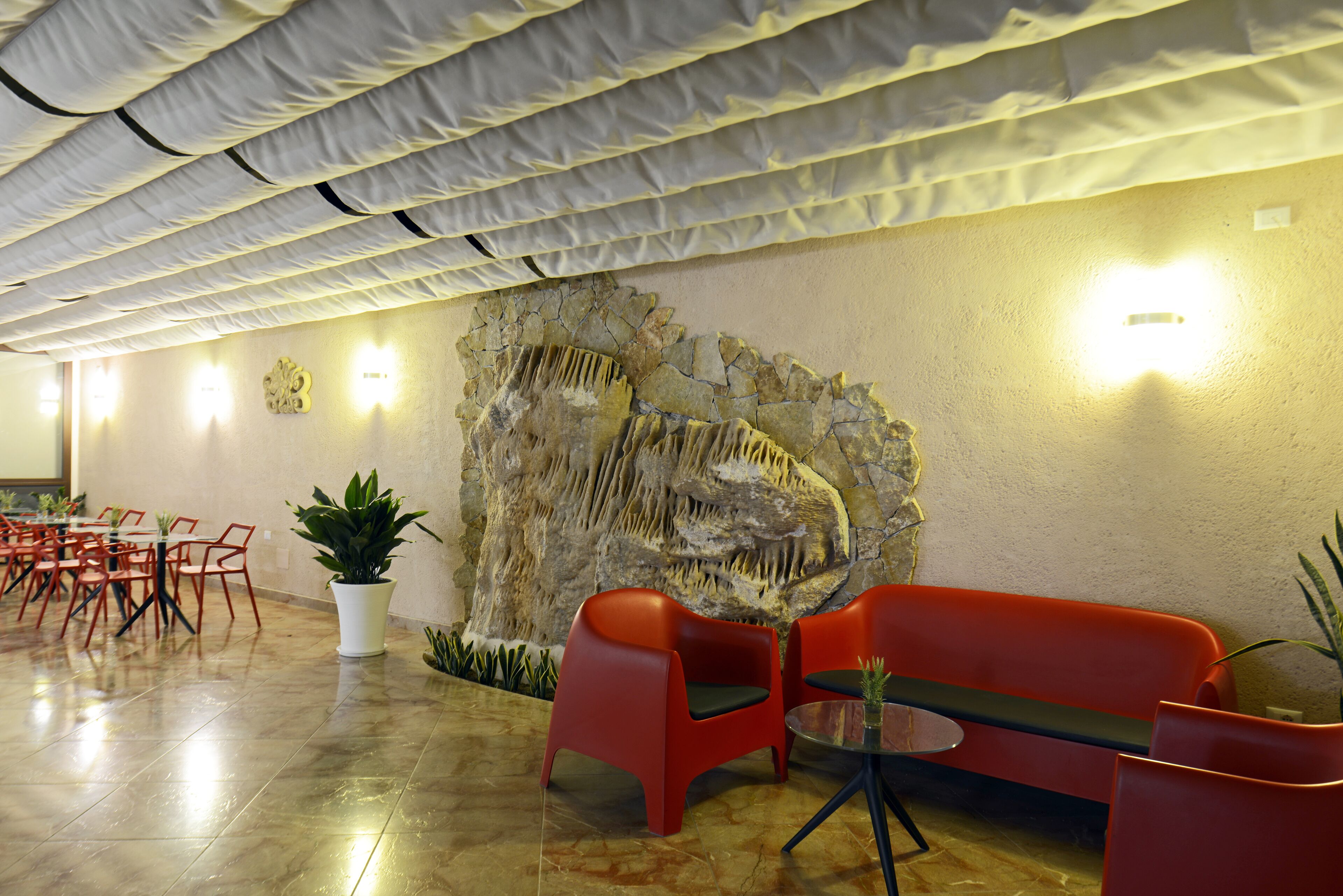 A modern interior with a stone wall, red chairs, a glass table, and a ceiling with fabric panels.