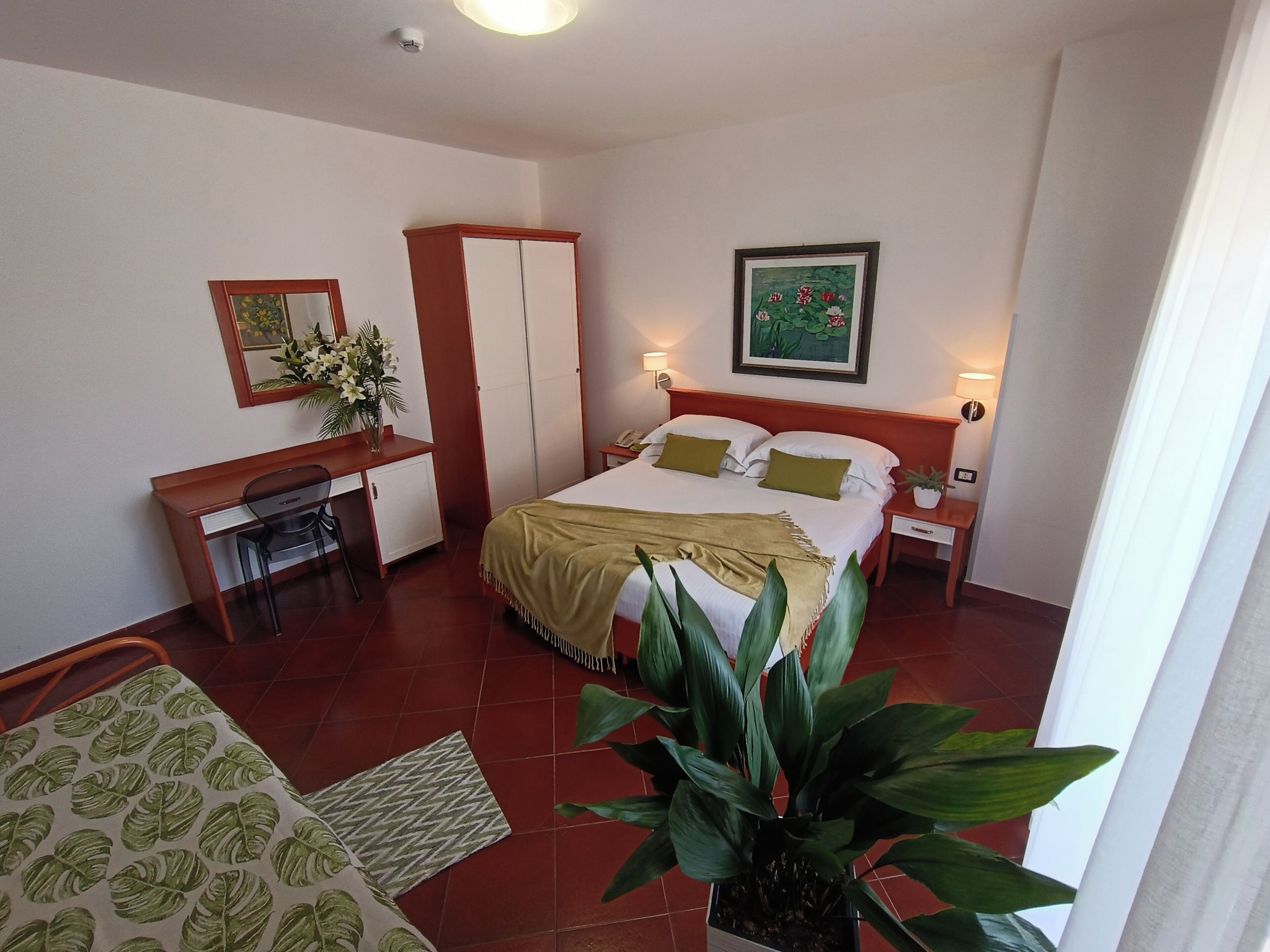 Habitación de hotel con cama, escritorio, silla, mesita de noche y una planta grande en maceta.