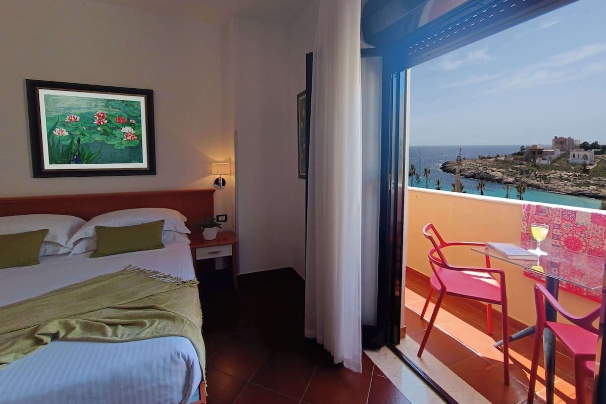 Ein Hotelzimmer mit Bett, Nachttisch, Lampe, einem gerahmten Bild und einem Balkon mit Tisch und Stühlen.