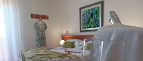 A hotel room with a neatly made bed, a bedside table with a glass of water, a painting on the wall, and a dress on a hanger.