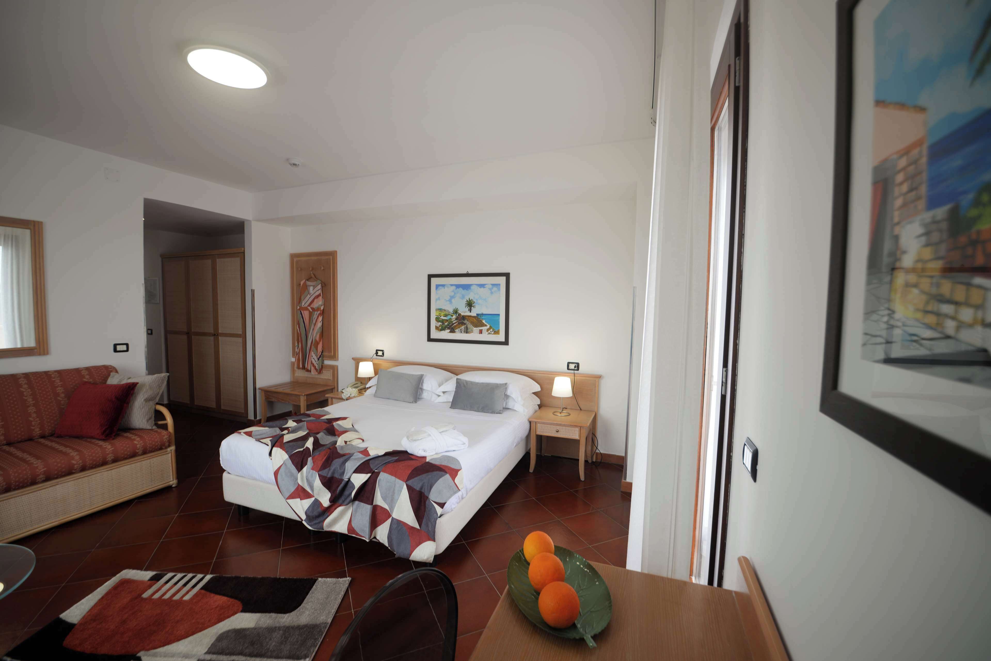 A hotel room with a bed, a sofa, a dining table with oranges, and a painting on the wall.