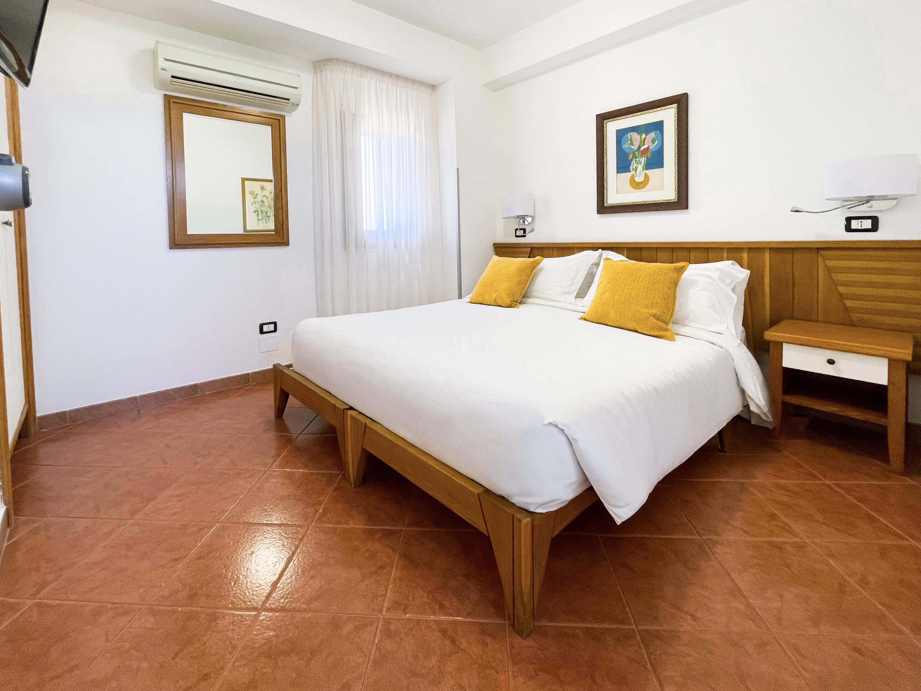 A hotel room with a bed, a nightstand, a wall-mounted TV, an air conditioning unit, a window with curtains, and a framed picture on the wall.